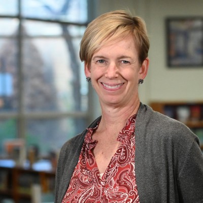 Alumna Erin Wyatt, PhD, MLIS, is pictured in a library setting. Erin has short, blonde hair and is wearing a red patterned shirt and a gray sweater.