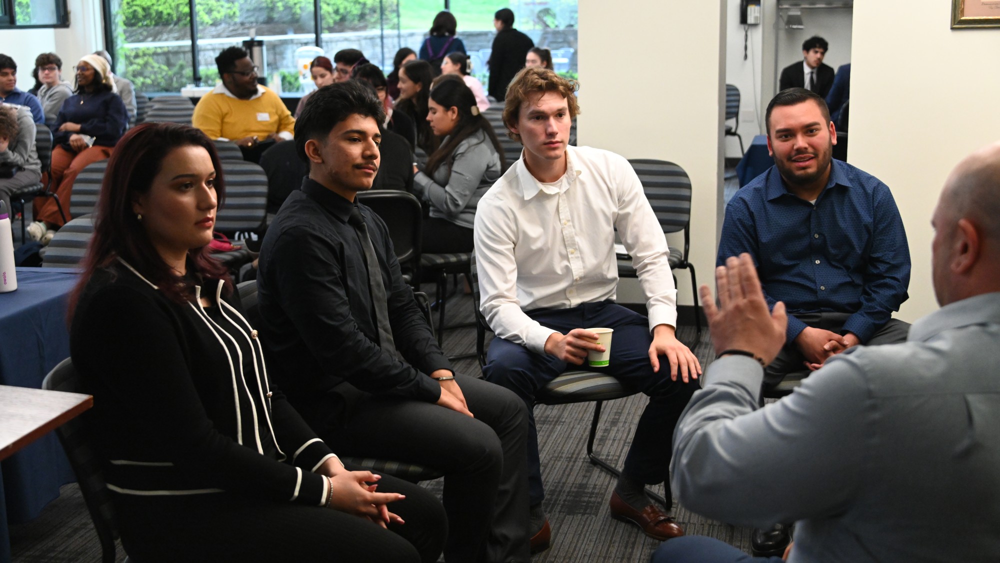 A group four university students, one female and three male, and one older male professor sit in a semi-circle engaged in a focused conversation in a professional setting. The younger participants are dressed in business casual attire, and one holds a cup. In the background, other small groups of students and professors are also interacting, suggesting a workshop or networking event. The atmosphere appears collaborative and attentive.