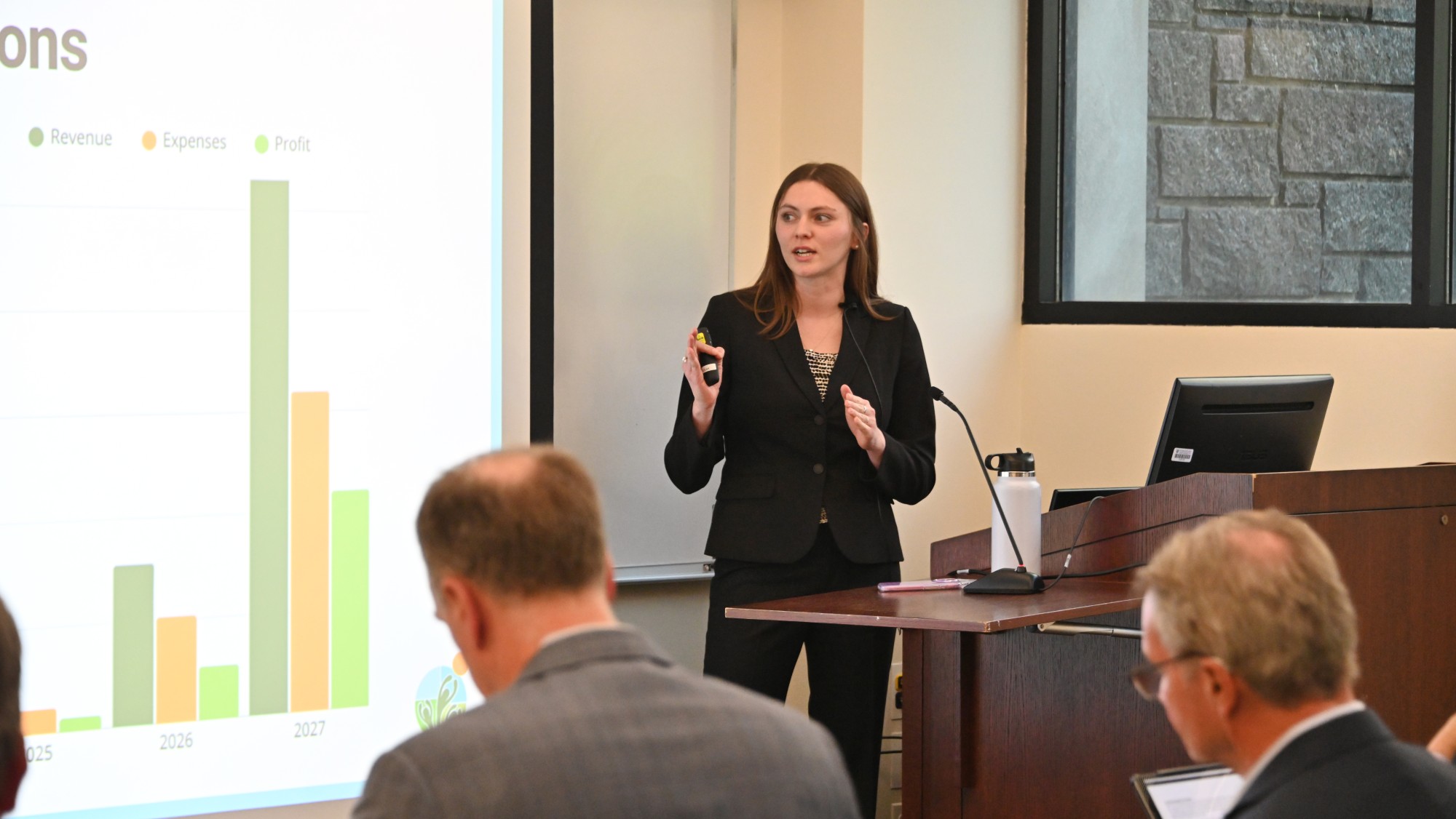 A woman in a suit presents a bar chart showing financial growth to a seated audience in a conference room.