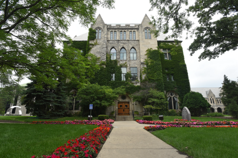 Lewis Hall shown in spring, with green ivy covering the building and red and purple flowers lining the walkways.