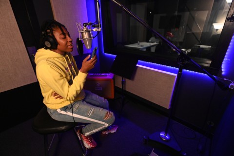 Dominican student Timiya Ray is wearing a yellow hoodie and ripped jeans sits on a stool in a dimly lit recording studio, singing into a large microphone. Timiya wears headphones and holds a phone, reading lyrics. The room is lit with blue LED lights and features soundproofing panels, a window to the control room, and recording equipment.