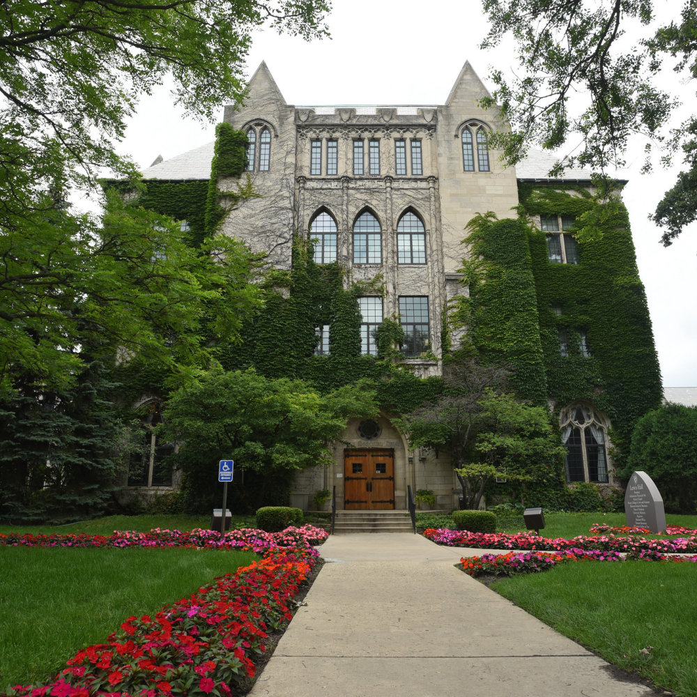 Lewis Hall shown in spring, with green ivy covering the building and red and purple flowers lining the walkways.