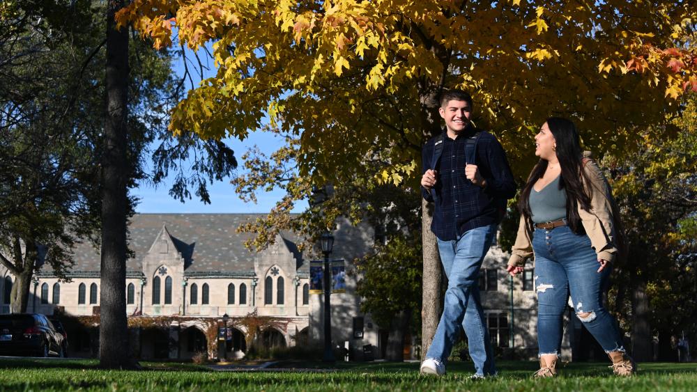 Two students wearing jeans and jackets walking across a field of grass on Dominican University's campus in front of Mazzuchelli Hall. . There is a tree with leaves turning yellow in the background.