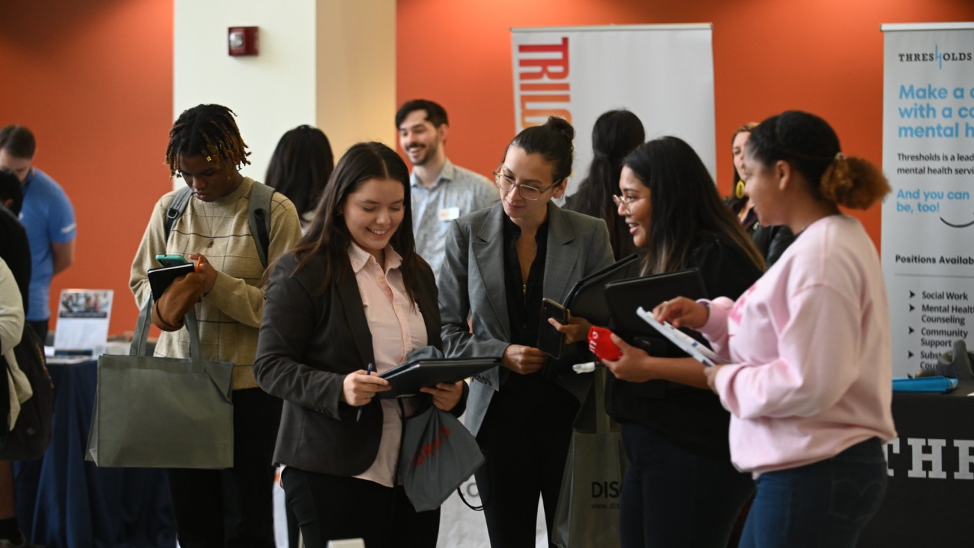 Four female students wearing business clothing are talking at Dominican University's Career Fair. One student is holding a folio in her hands, and the students are all looking down at the folio.