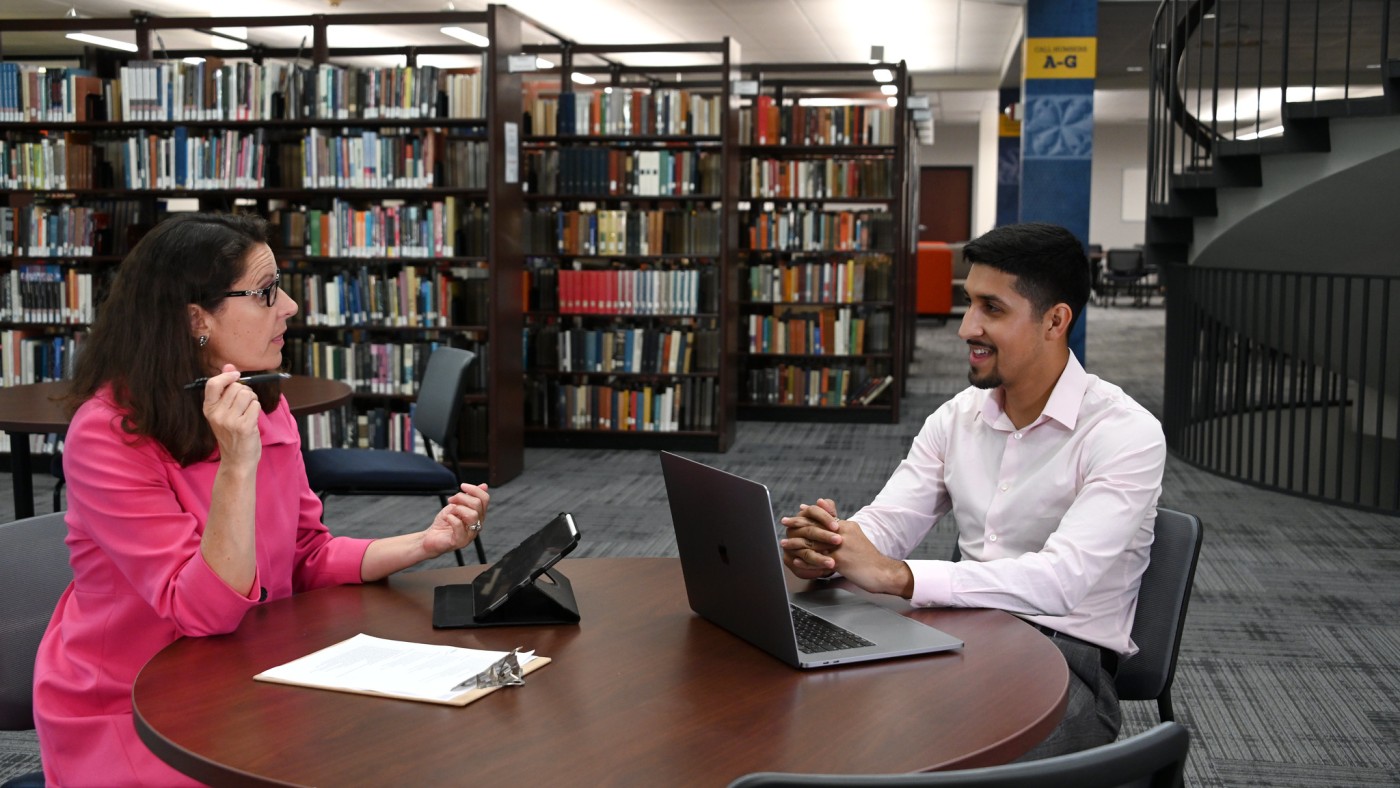 A female professor with dark curly hair and glasses, wearing a pink jacket, is seated at a table in front of a tabley. She is talking with a male student with dark hair, who is wearing a white shirt. He has a laptop in front of him. Library shelves with books are in the background.