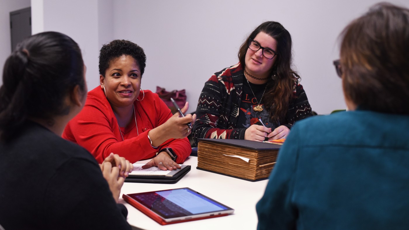  A graduate student in an information studies class speaks while others listen and take notes around a table. Tablets, notebooks, and a large reference book are visible, suggesting a collaborative discussion.