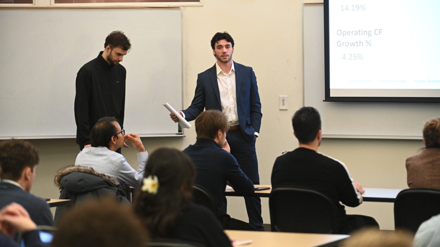  Two business students present in front of a classroom, one speaking while the other stands nearby. A slide displaying financial metrics is projected, and seated classmates listen attentively.