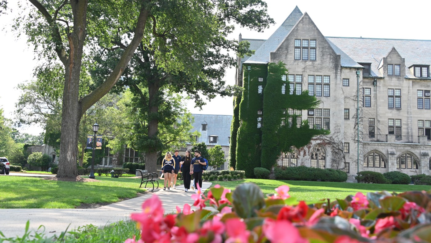  Four students walk along a tree-lined campus path past Lewis Hall, a historic, ivy-covered buildingon Dominican University's River Forest Campus, with bright pink flowers in the foreground.