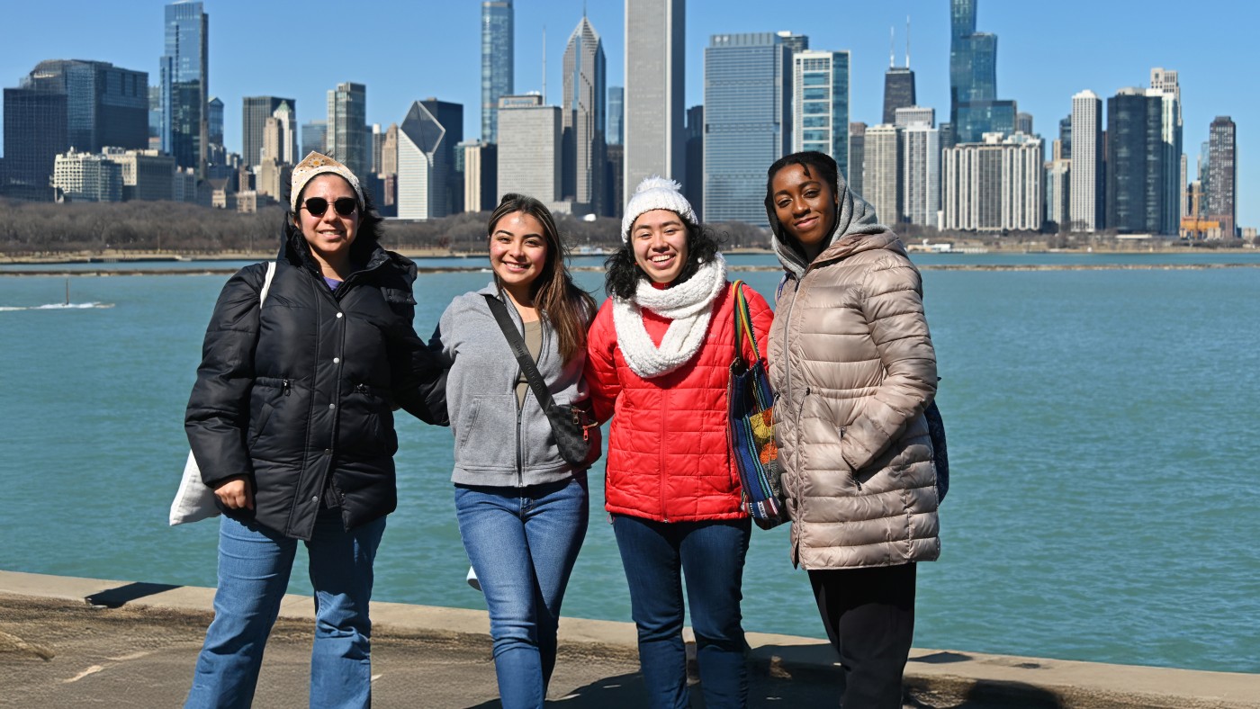 Four Dominican University students pose at the lakefront on a sunny day. They are wearing coats. The Chicago skyline is in the background.