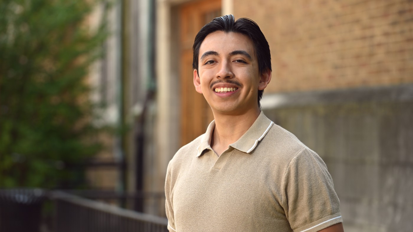 Student Aldo Cervantes stands outside of Dominican University's Lewis Hall on the River Forest Campus. He has short, dark hair and a mustache. He is smiling at the camera and wearing a tan polo shirt.