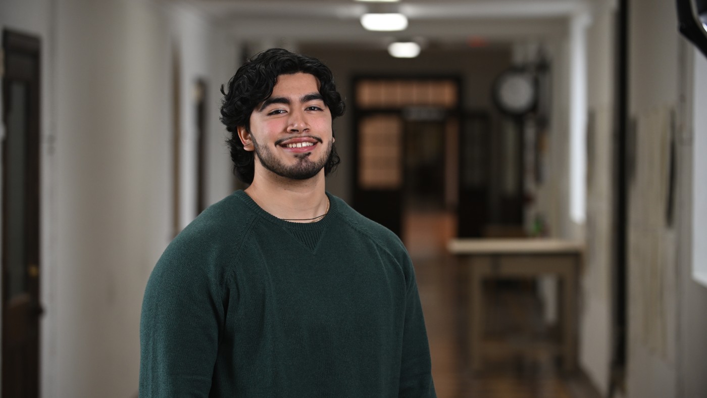 Marketing student Andy Nunez stands in a hallway at Dominican University's River Forest Campus. He has dark hair and is smiling and wearing a dark green sweater.