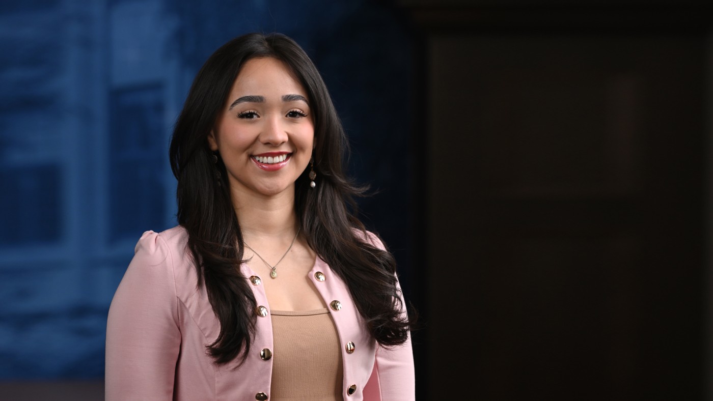 Student Jocelyn Ramirez stands in front of a graphic of Dominican University's River Forest campus. She has long, dark hair and is smiling. She is wearing a tan shirt and a light pink jacket.