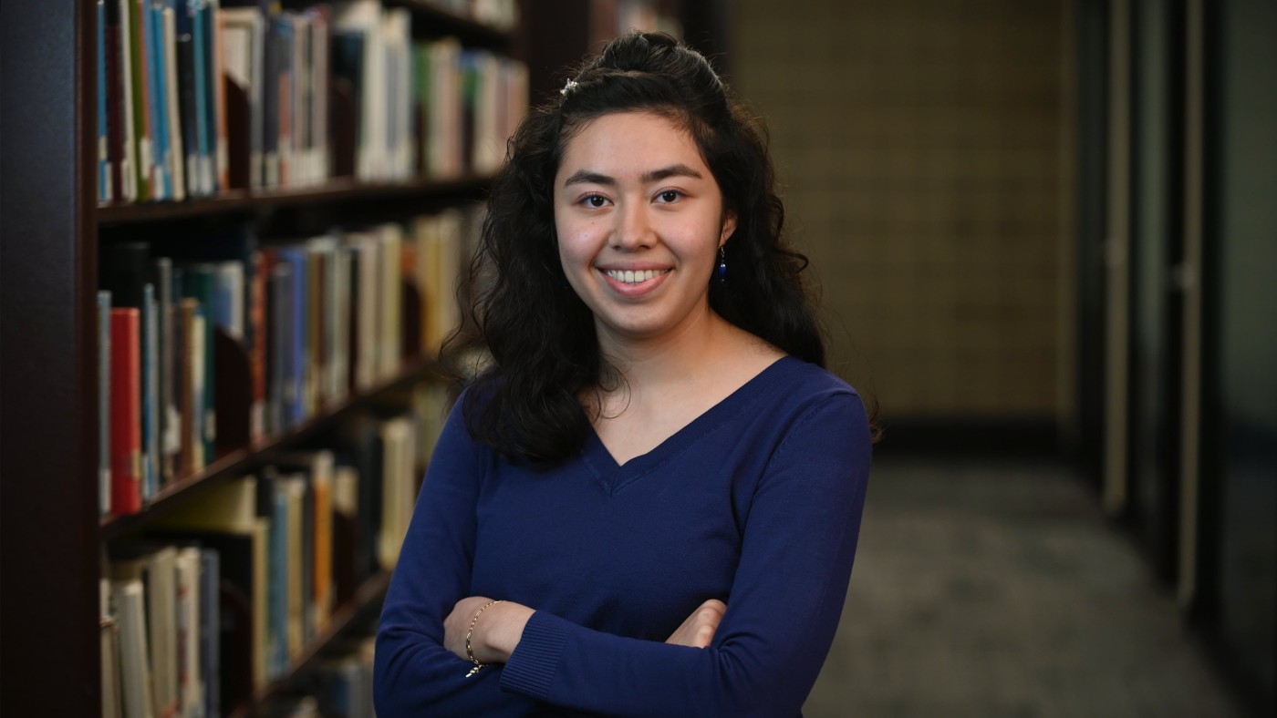 Student Isabella Flores is standing in the library in front of bookshelves. She has long, dark curly hair, is smiling and has her arms folded in front of her. She is wearing a dark blue shirt. a