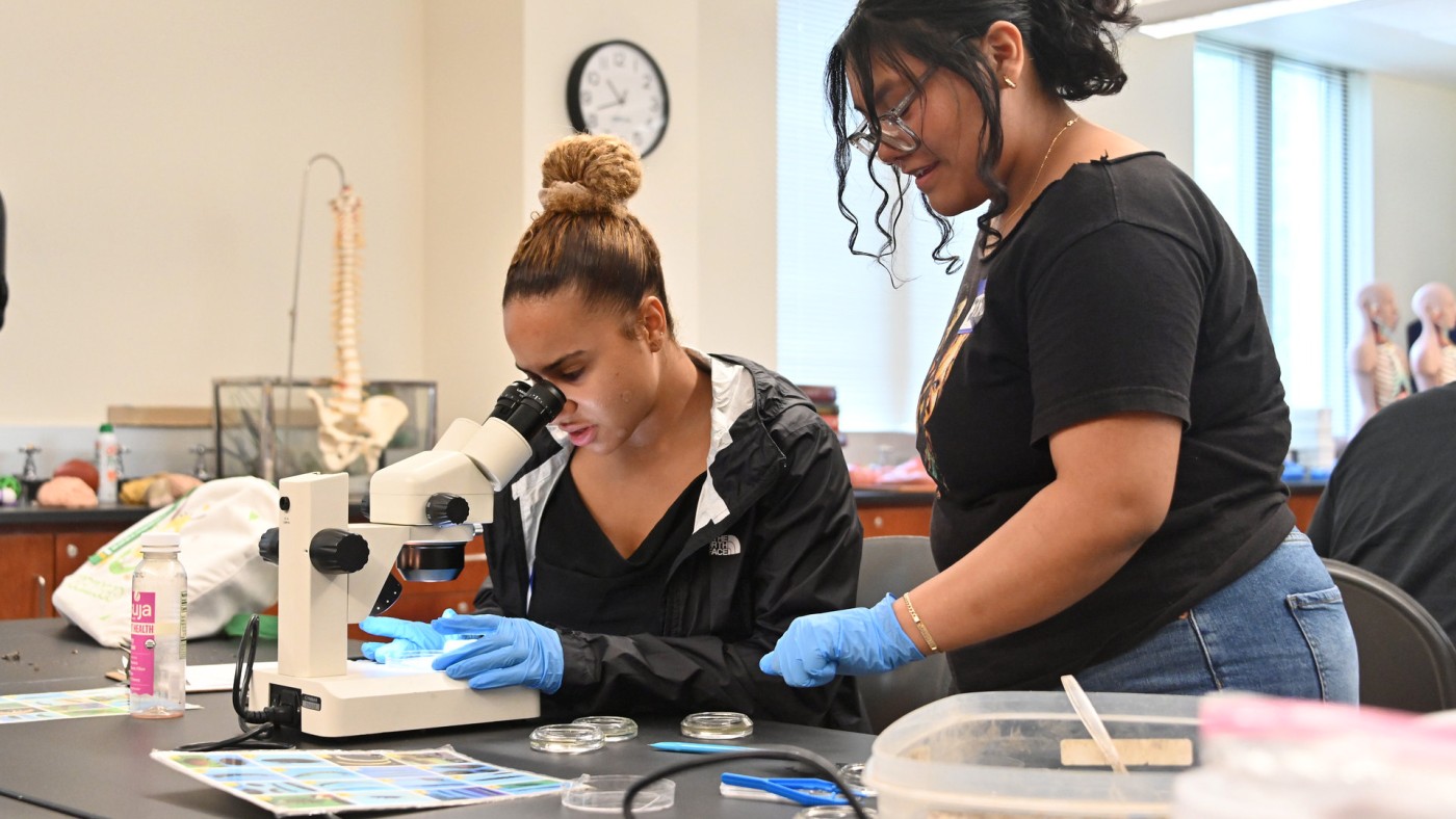 Two female students in a science lab, one looking through a microscope.
