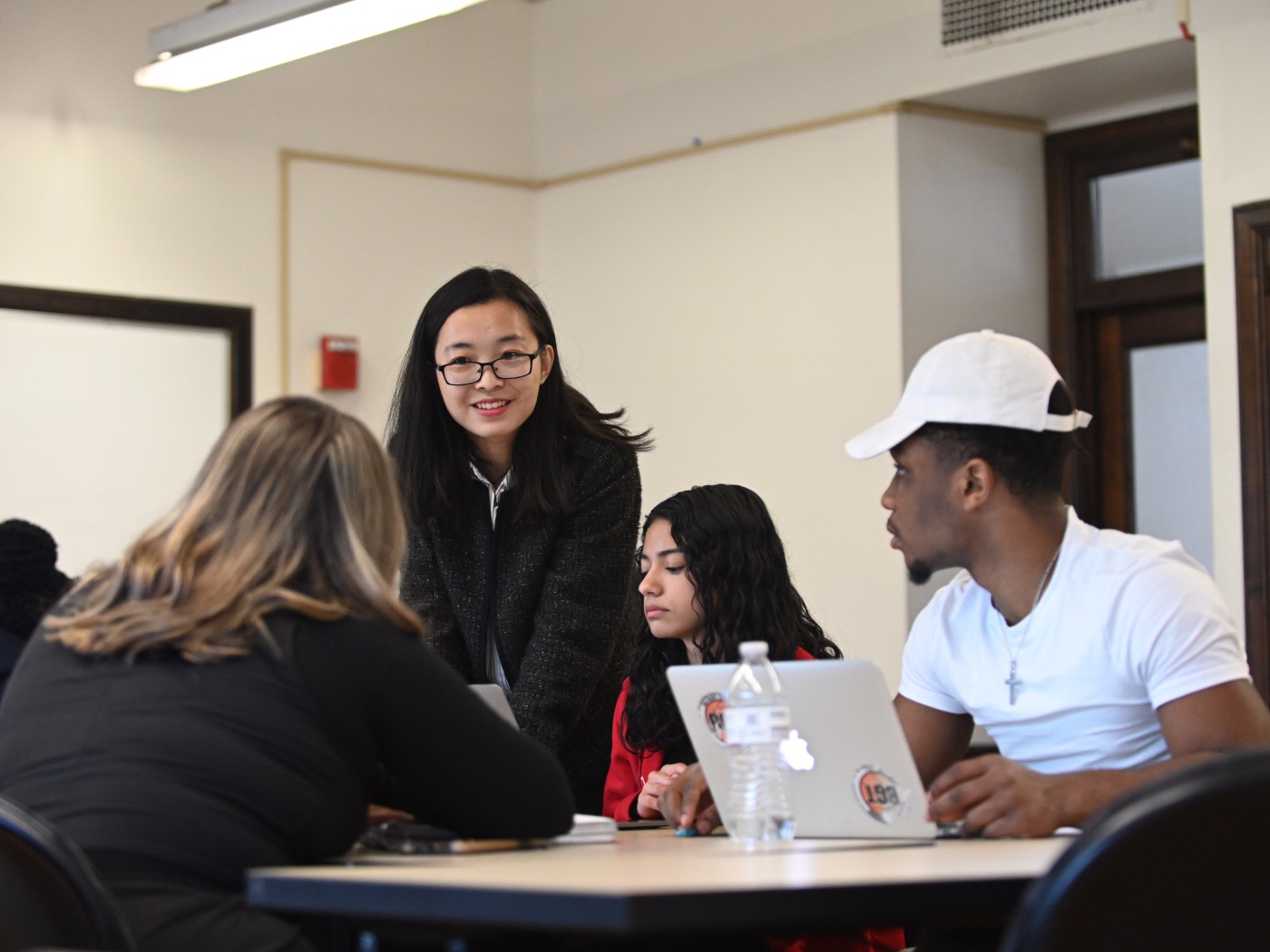  A business professor leans over a table to speak with three students working on laptops during class.