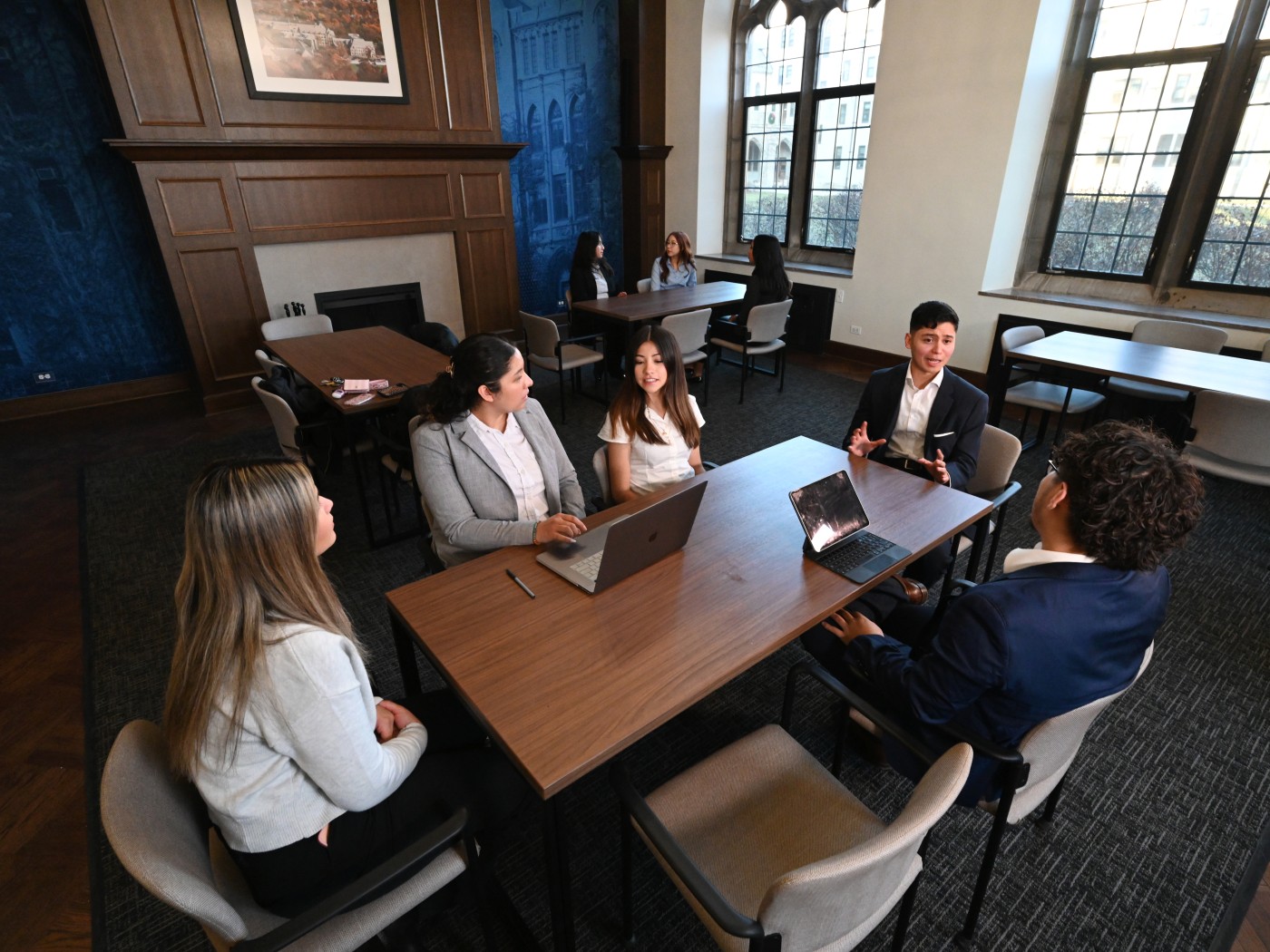  Students in business attire sit around a table in an academic study room, discussing with laptops open. Other small groups talk in the background.