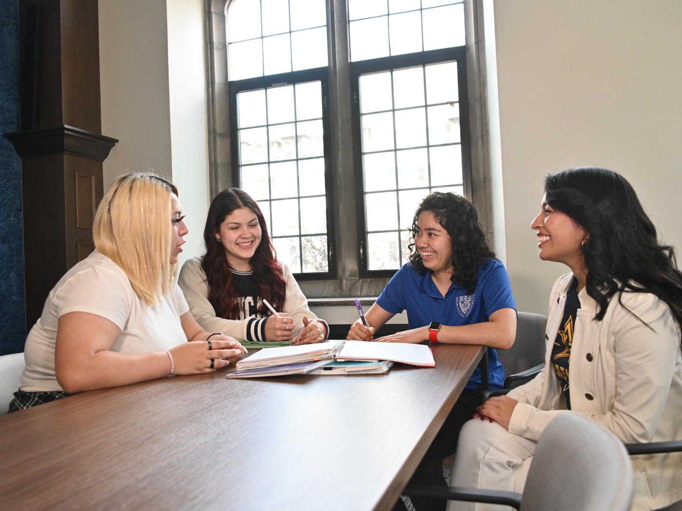 Four female Dominican University students sit around a table in a classroom, talking and taking notes.